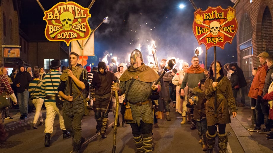 Members of the Battel Bonfire Boyes in Saxon costume and chain mail carrying flaming torches and two red-and-yellow skull-and-crossbones society banners along Battle High Street at night, with spectators on the pavements