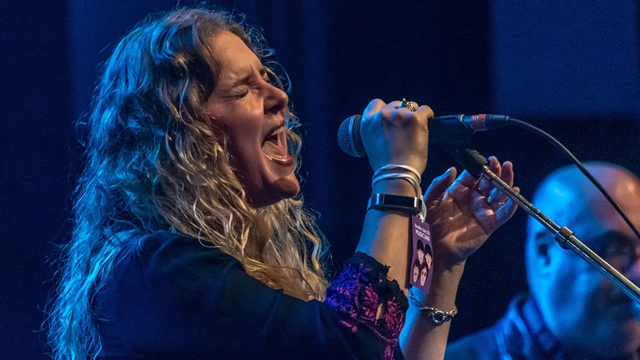A singer with long curly hair sings passionately into a microphone under blue stage lights at Hastings Beatles Day, with a musician pass hanging from her wrist