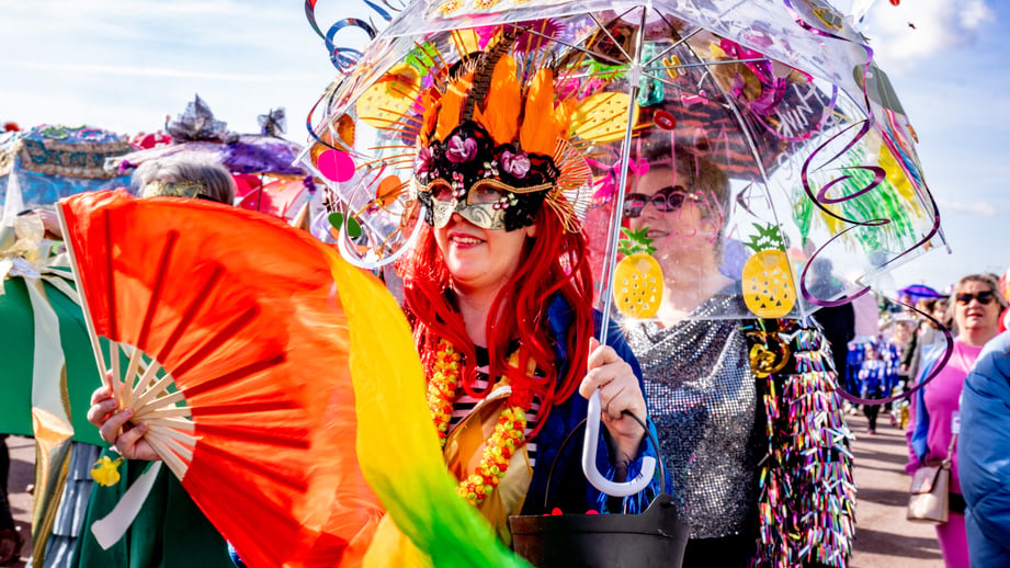 A woman in a feathered Mardi Gras mask and red wig carries a decorated clear umbrella in the Hastings Fat Tuesday umbrella parade, with a rainbow fan in the foreground and costumed paraders behind under a blue sky