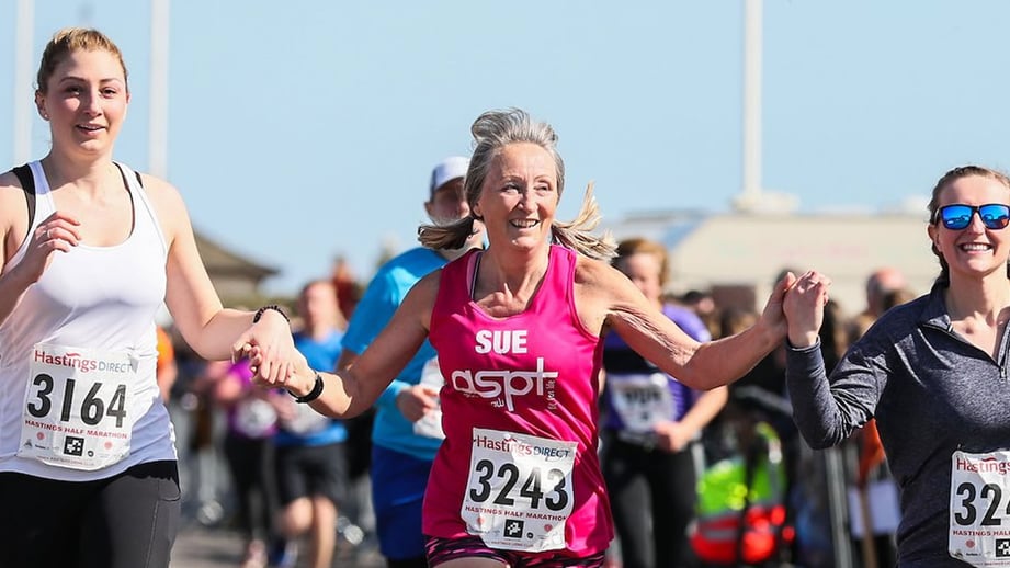 Three smiling women in Hastings Half Marathon race numbers run hand in hand along the seafront, with spectators behind crowd barriers