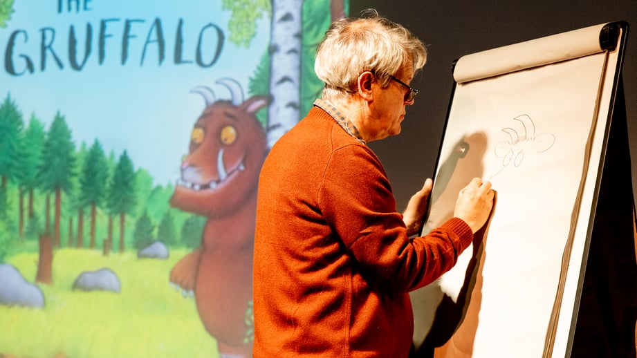 Illustrator Axel Scheffler sketching on a flip chart on stage, with a projected Gruffalo picture-book cover behind him