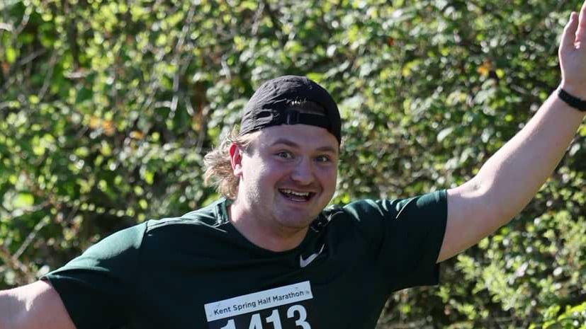 A grinning runner in a backwards cap and Kent Spring Half Marathon race number raises an arm as he passes a hedge