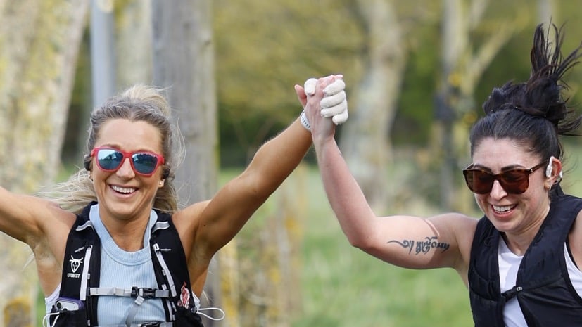 Two smiling runners in sunglasses and race vests hold hands aloft as they run along a tree-lined path
