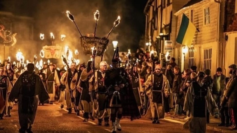 Robertsbridge Bonfire Society members in monks' habits and a kilted piper lead a torchlit procession down a village street at night, under the society's banner, past spectators and weatherboarded houses